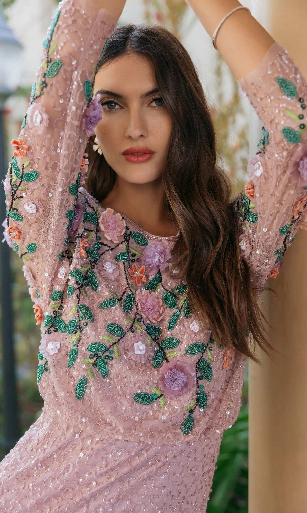 A woman with long brown hair stands with her arms raised, looking at the camera, wearing the Mac Duggal 9341 long formal dress—an elegant pink gown adorned with floral embroidery and sequin embellishments.